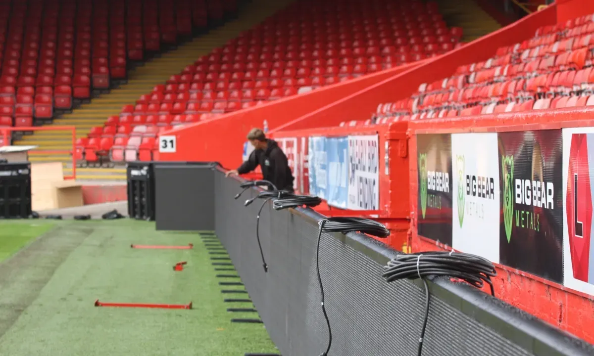 A technician fitting the LED boarding at Pittodrie Stadium, cabinets and cables along the pitch side