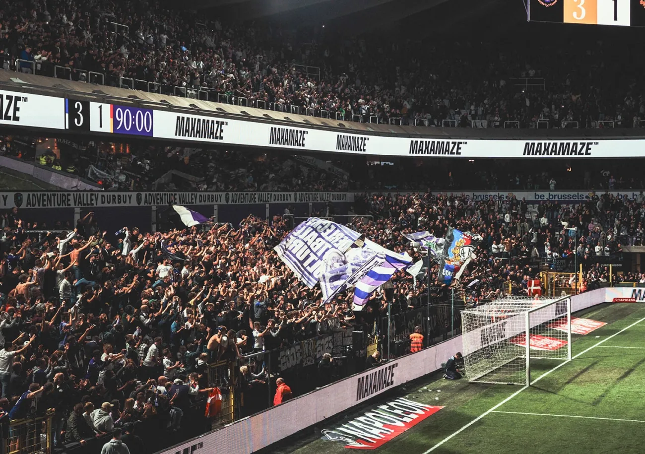 Lotto Park on a matchday: a packed stand behind the lit LED perimeter, the scoreboard reading 3-1 at 90 minutes